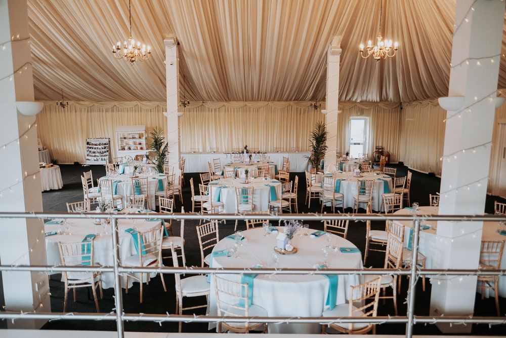 Elegant wedding reception venue with draped fabric ceiling, crystal chandeliers, and tables set in white and turquoise decor.