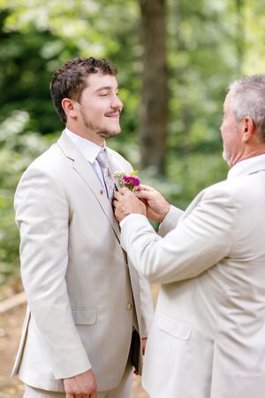 A boutonniere being pinned onto a light-colored suit jacket during wedding preparations.