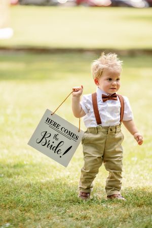 A toddler in suspenders and bow tie holds a 'Here Comes the Bride' sign while standing on grass.