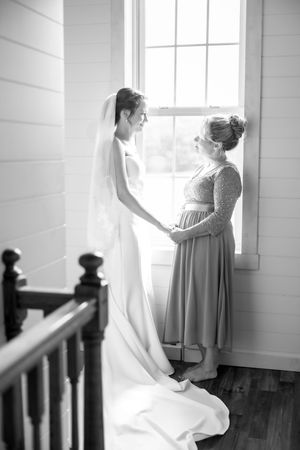 Mother and daughter share an intimate moment by a window in elegant wedding attire.