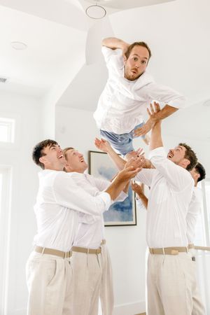 Groomsmen in white shirts playfully tossing someone in the air indoors.