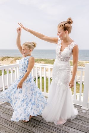 A bride in white gown dances with young girl in blue floral dress on oceanfront deck.
