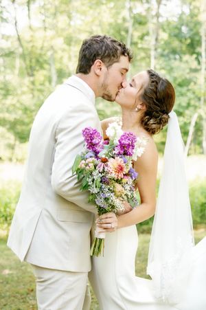 Newlyweds share a kiss while holding a vibrant purple and pink wedding bouquet.