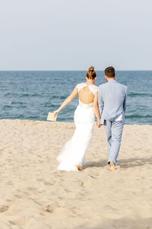 Newlyweds walk hand in hand along beach with ocean in background.