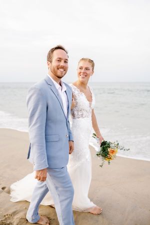 Newlyweds walk hand in hand along beach in light blue suit and white dress.
