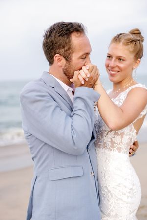 Newlywed couple shares intimate moments on a cloudy beach, with the bride in white lace and groom in light blue suit.