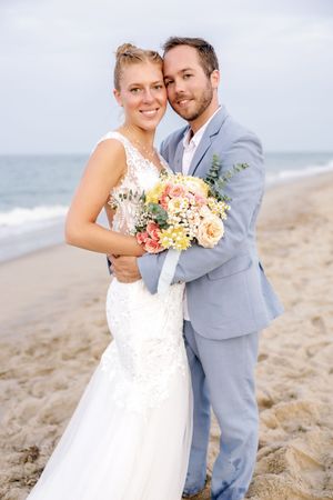Wedding couple poses romantically on sandy beach with coral and cream floral bouquet.