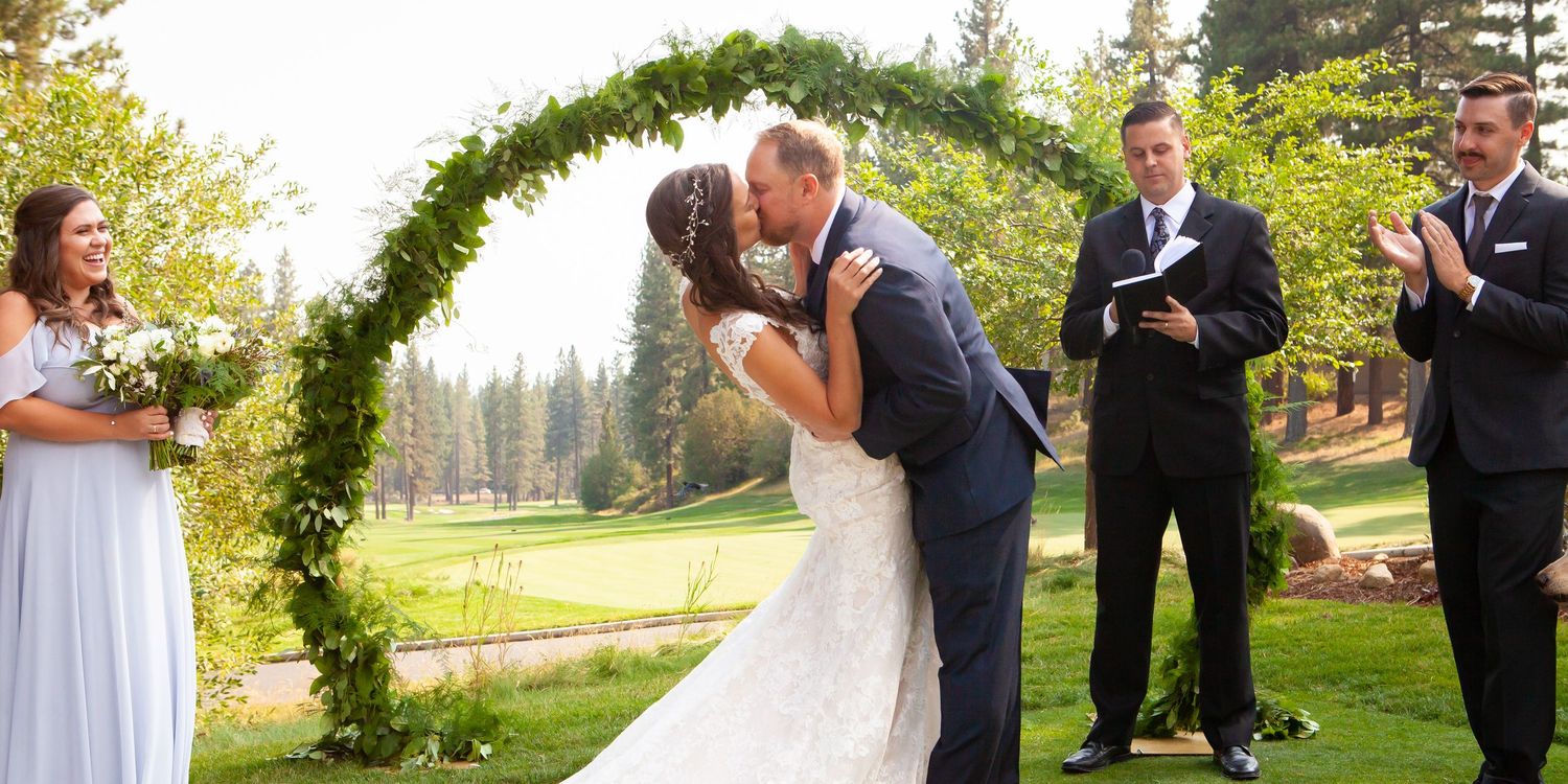 A bride and groom share a romantic kiss during their outdoor wedding ceremony surrounded by greenery and guests.