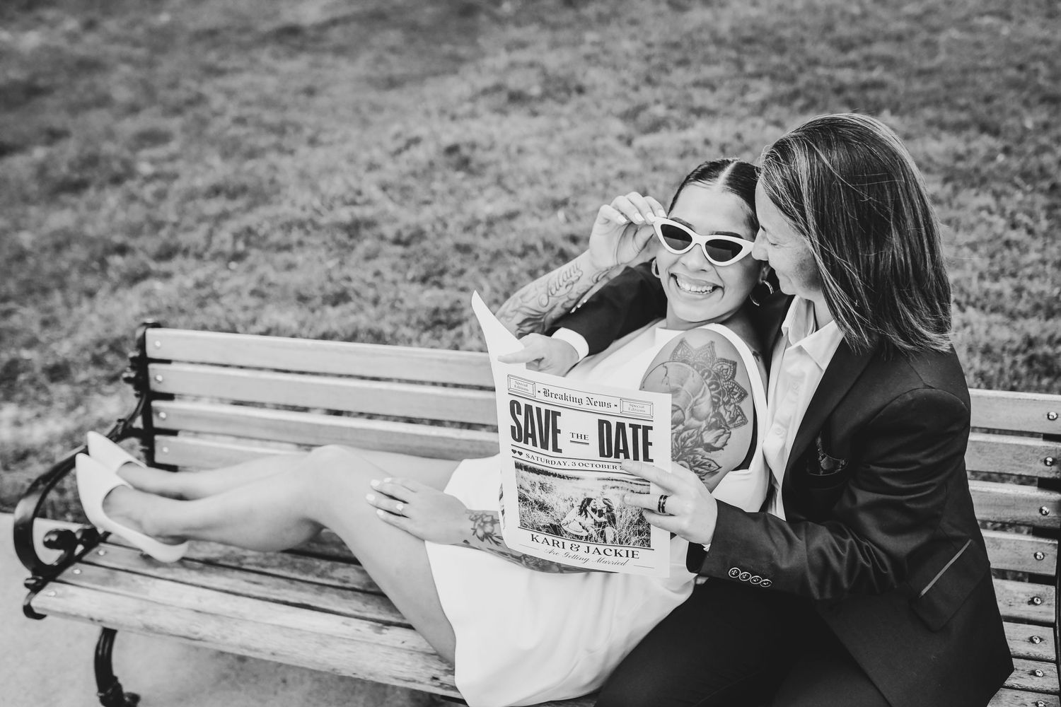 High-fashion engagement photo of a same-sex couple reading their save-the-date magazine on a park bench, captured in an editorial style

Sli