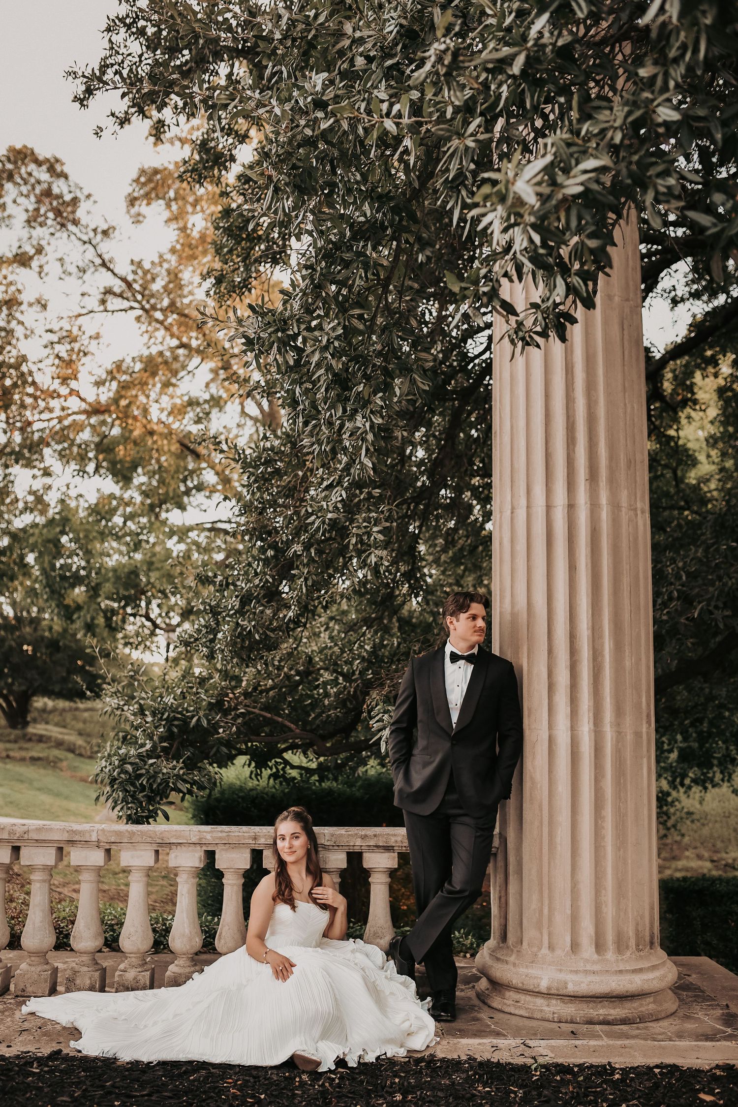 Bride in a white gown sits on stone steps while the groom in a black tuxedo leans against a tall column in a lush garden.