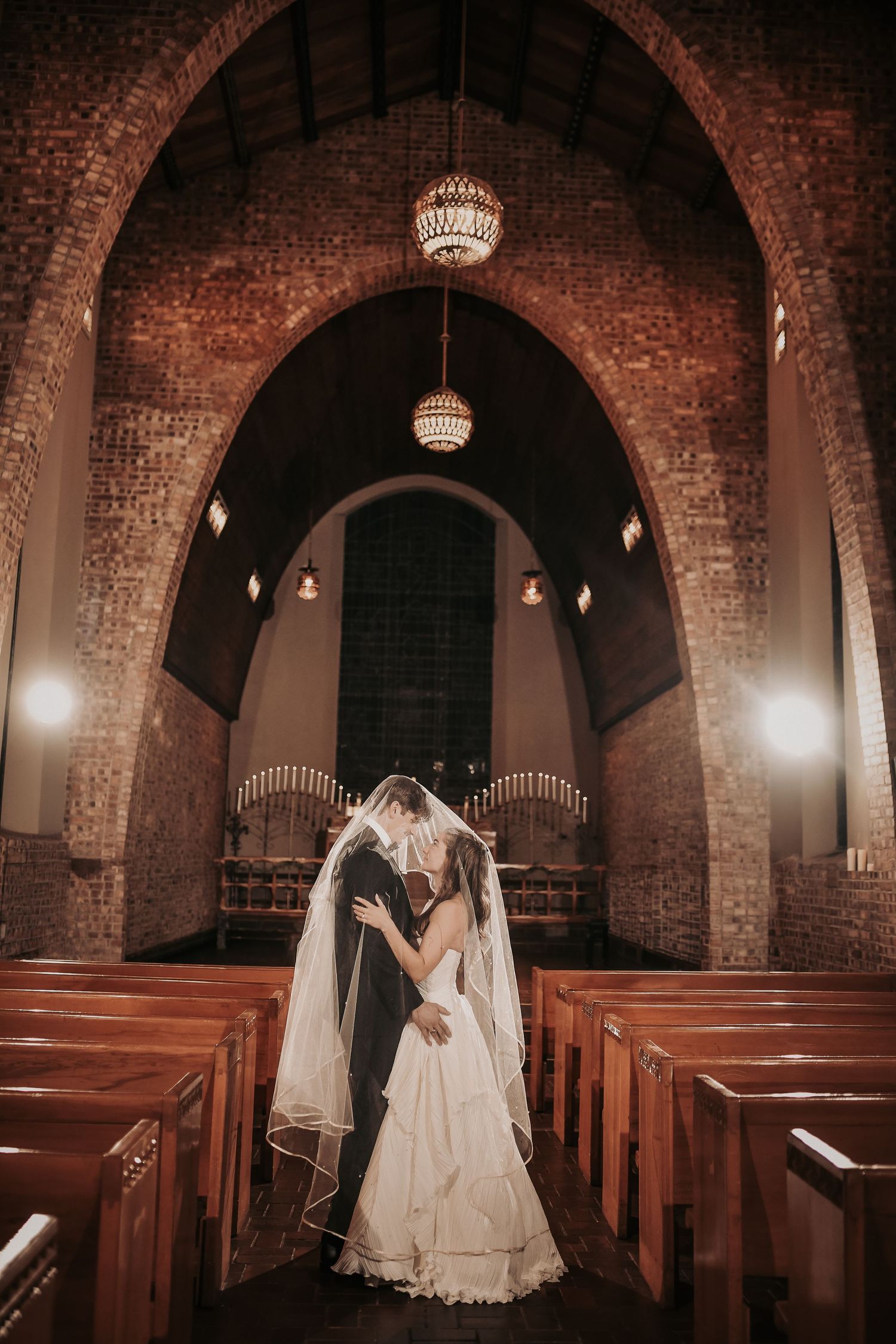 Bride and groom embrace under a sheer veil in the aisle of a historic brick chapel with wooden pews and glowing pendant lights.