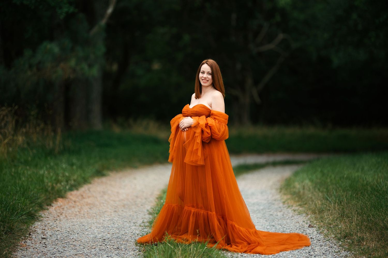 A flowing burnt orange gown with sheer fabric trails elegantly along a rustic dirt path through green foliage. Maternity Hendersonville