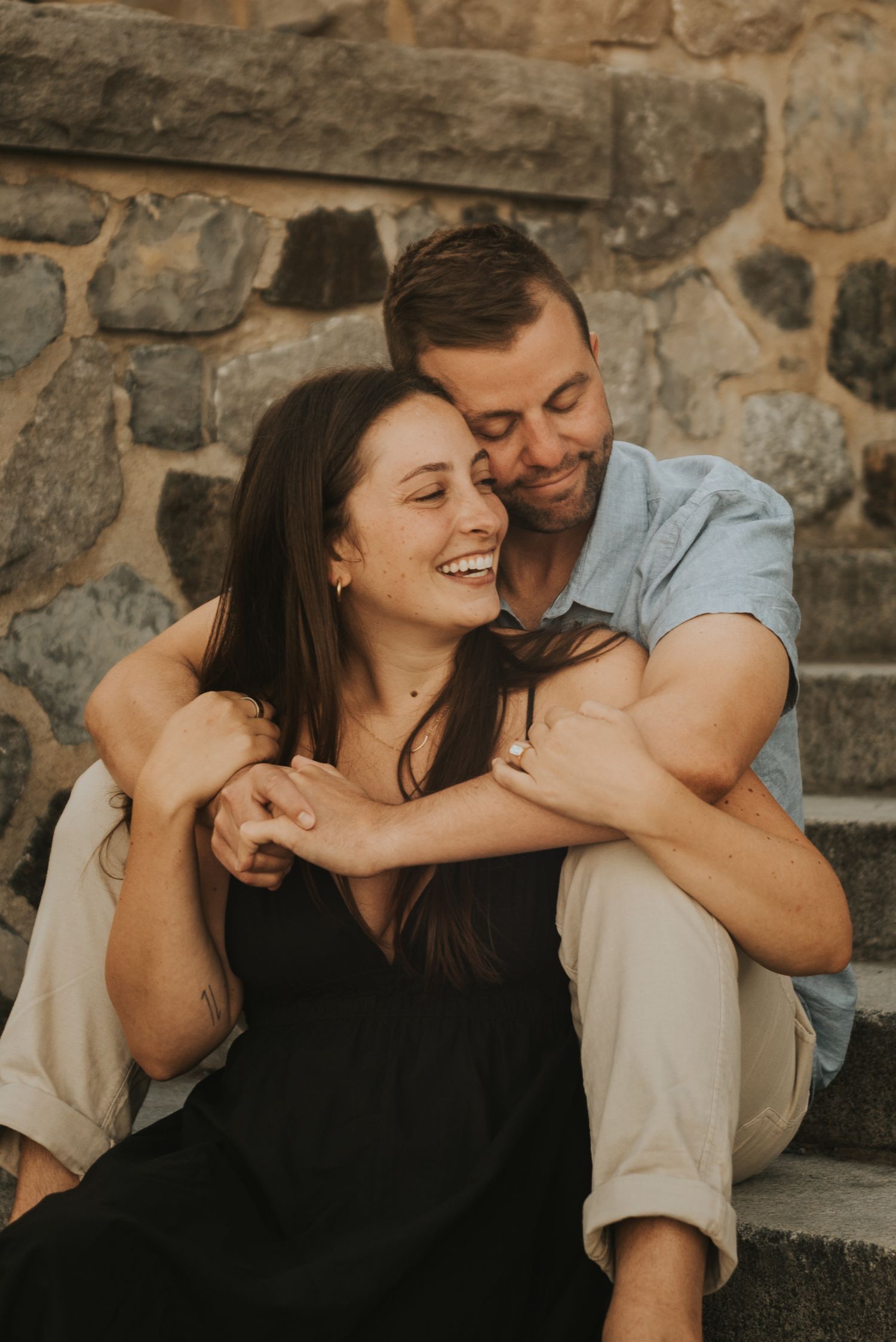 A couple shares a joyful moment while sitting on stone steps during a casual outdoor photoshoot.