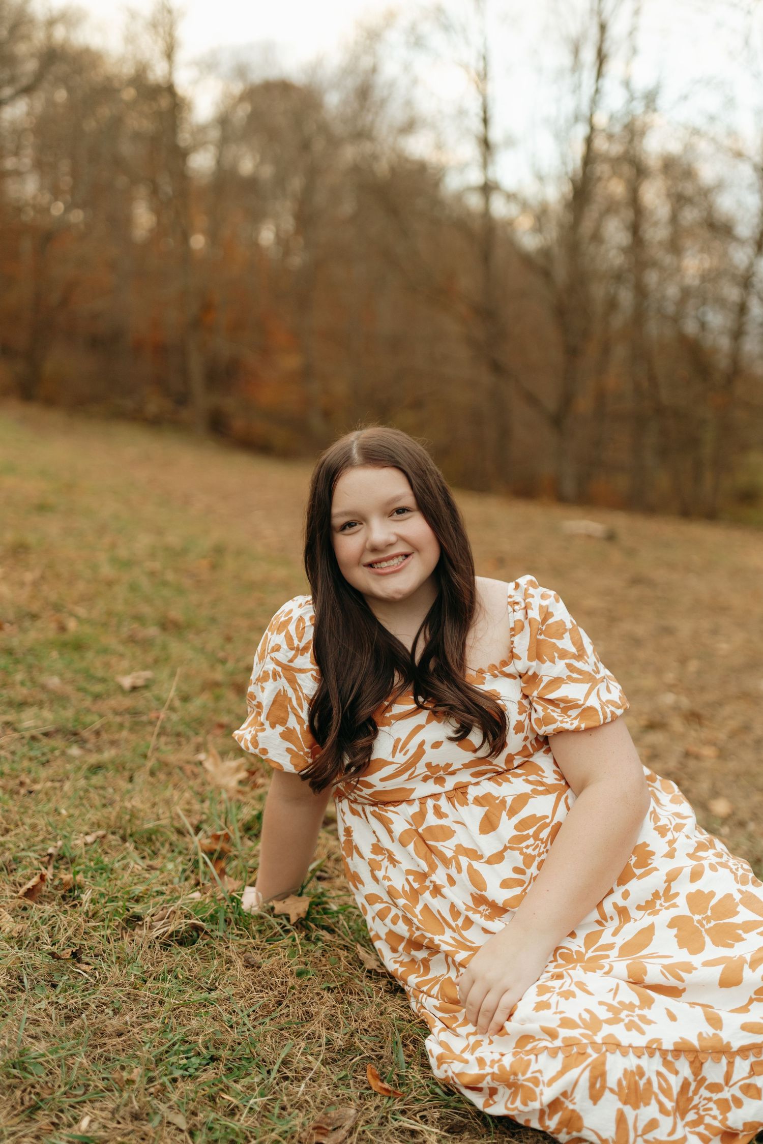 A person wearing a floral dress poses in a wooded area near a stream during autumn.