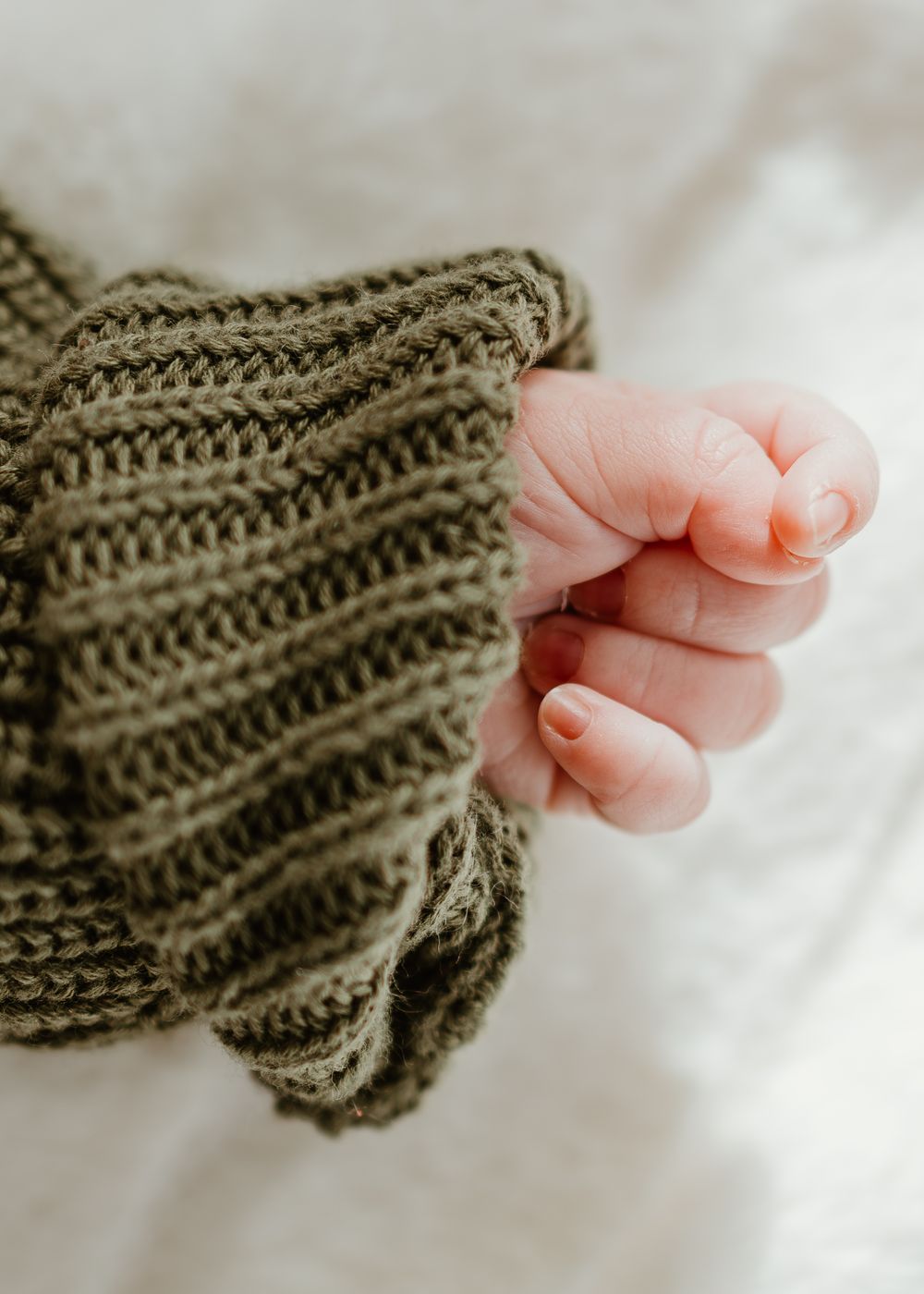 A tiny baby hand grasps the edge of an olive green knitted sweater against a white background.
