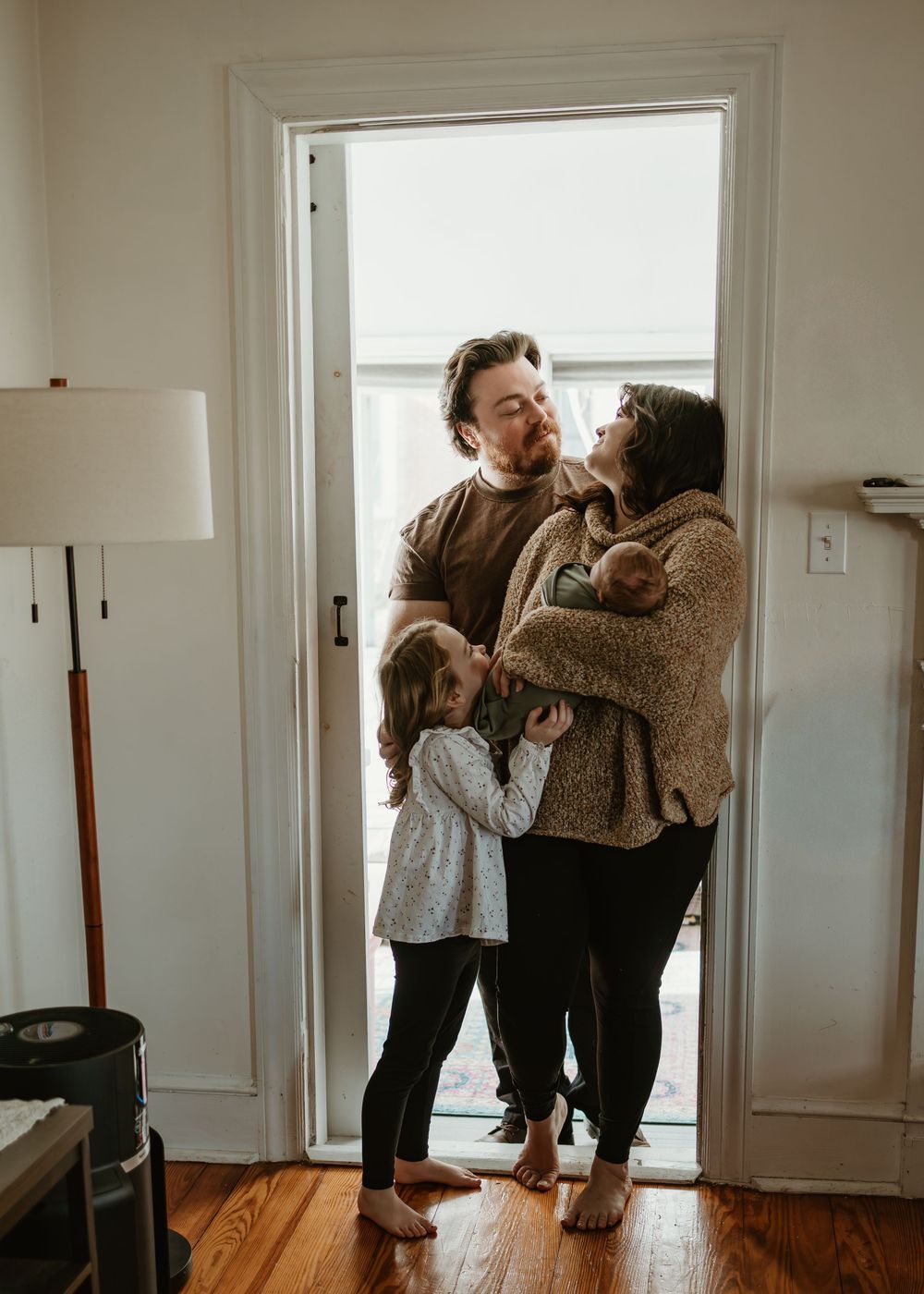 A parent and child share a tender moment in a sunlit hallway with hardwood floors.
