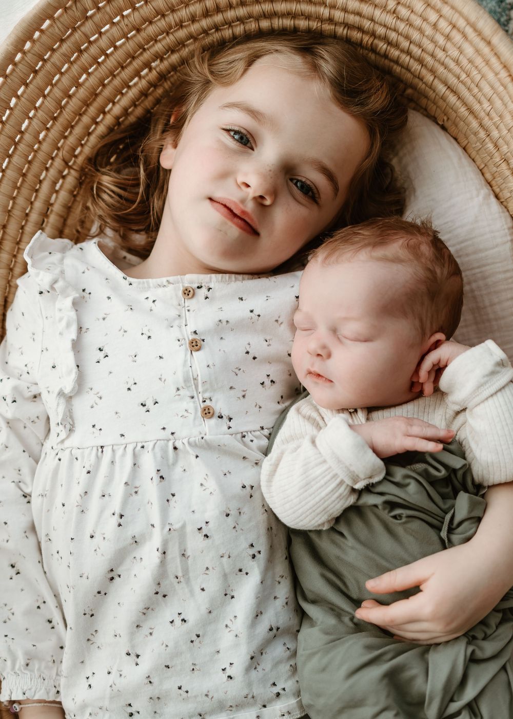 Two siblings cuddle together, with the older one wearing a white polka dot dress and the younger in green sleepwear.
