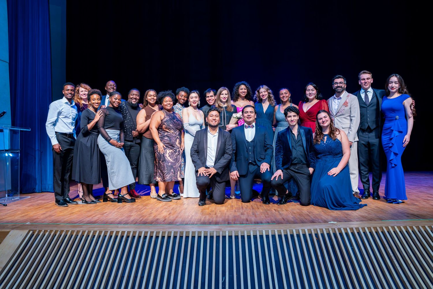Large theater ensemble poses together on stage for a group photo with blue lighting.