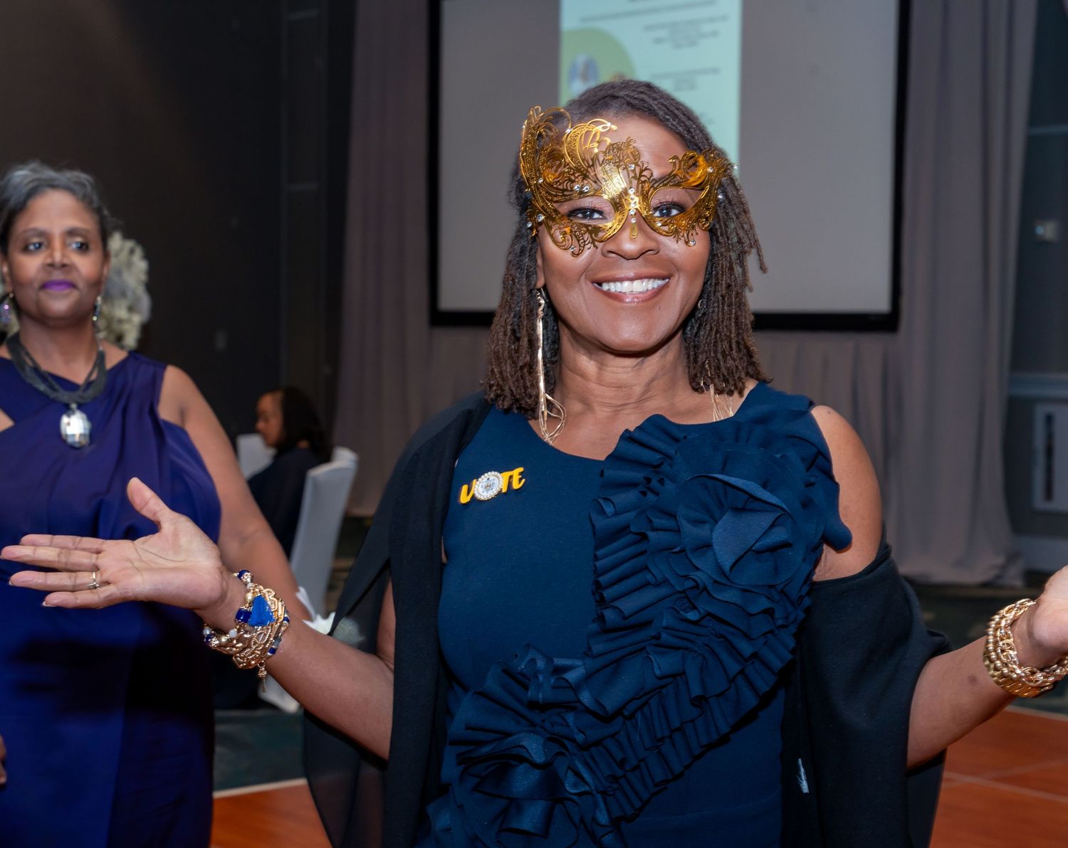 Woman in navy dress and gold masquerade mask smiles while dancing at formal event.