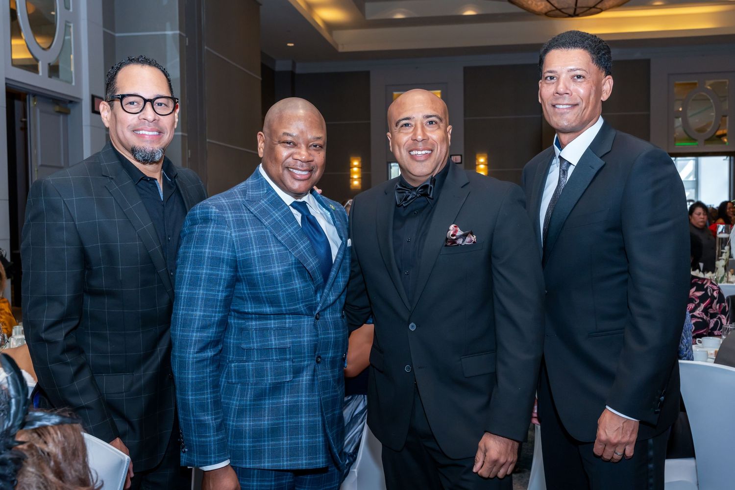 Four men in business suits pose together at formal gathering with elegant lighting.
