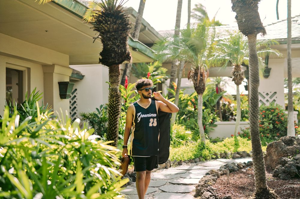 Person in a dark basketball jersey walks along a tropical resort path lined with palm trees and lush greenery.