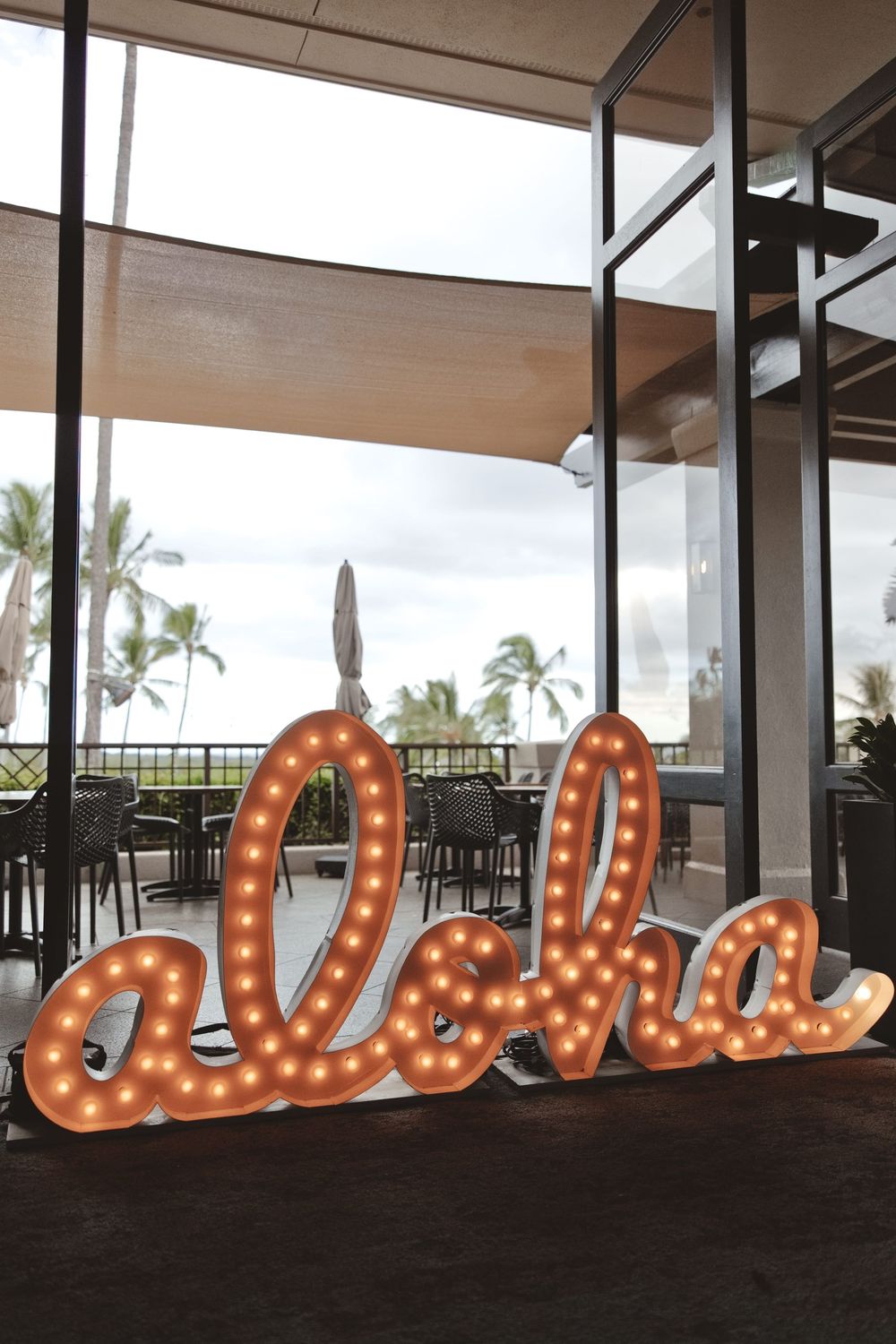 Large illuminated marquee letters spelling out a word in orange lights against a modern indoor-outdoor venue with palm trees.