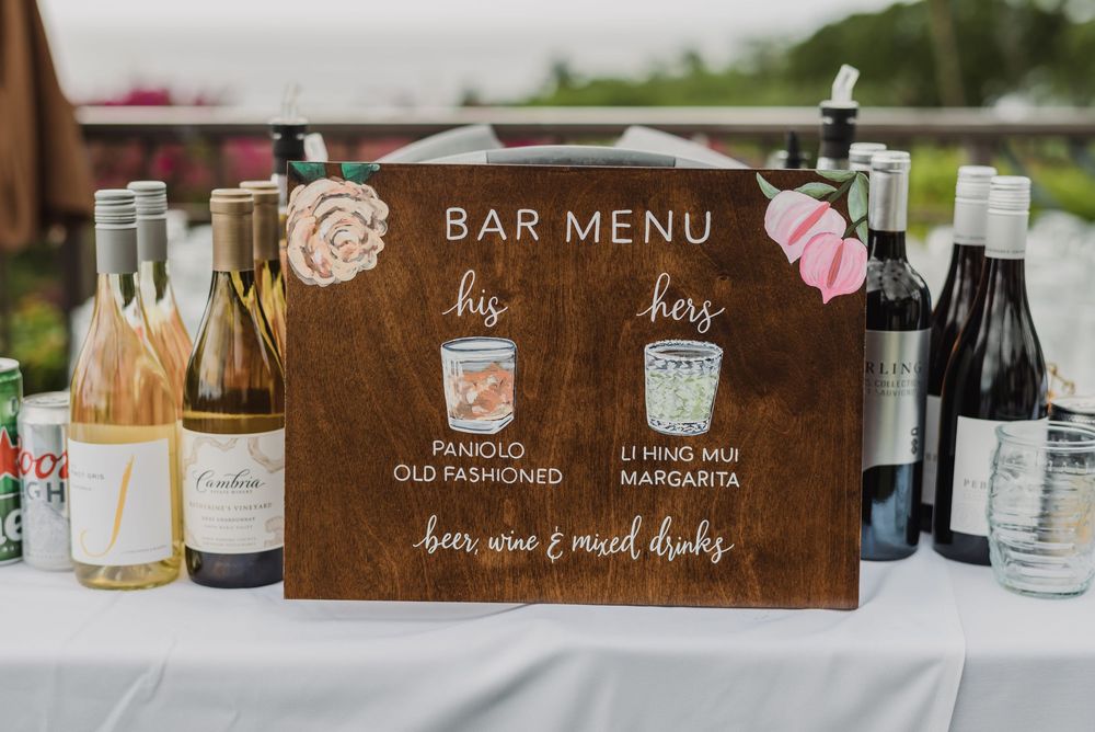 A rustic wooden bar menu sign displays cocktail options alongside assorted wine and liquor bottles on a white tablecloth.