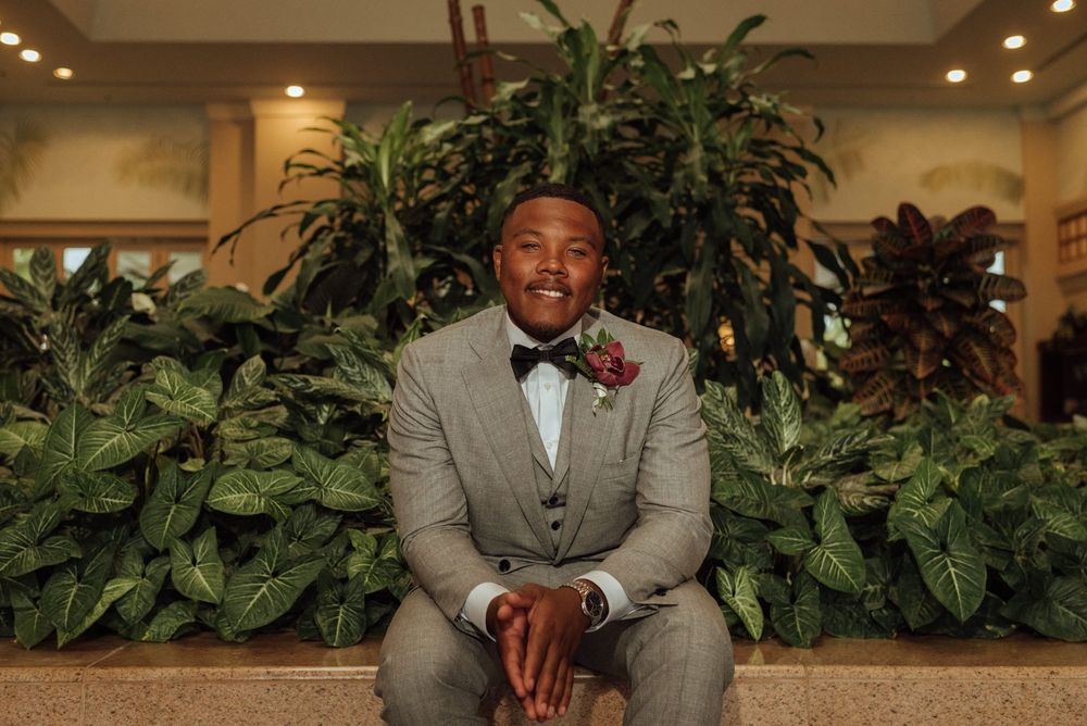 A groom in a gray suit poses in an elegant indoor setting with decorative plants.