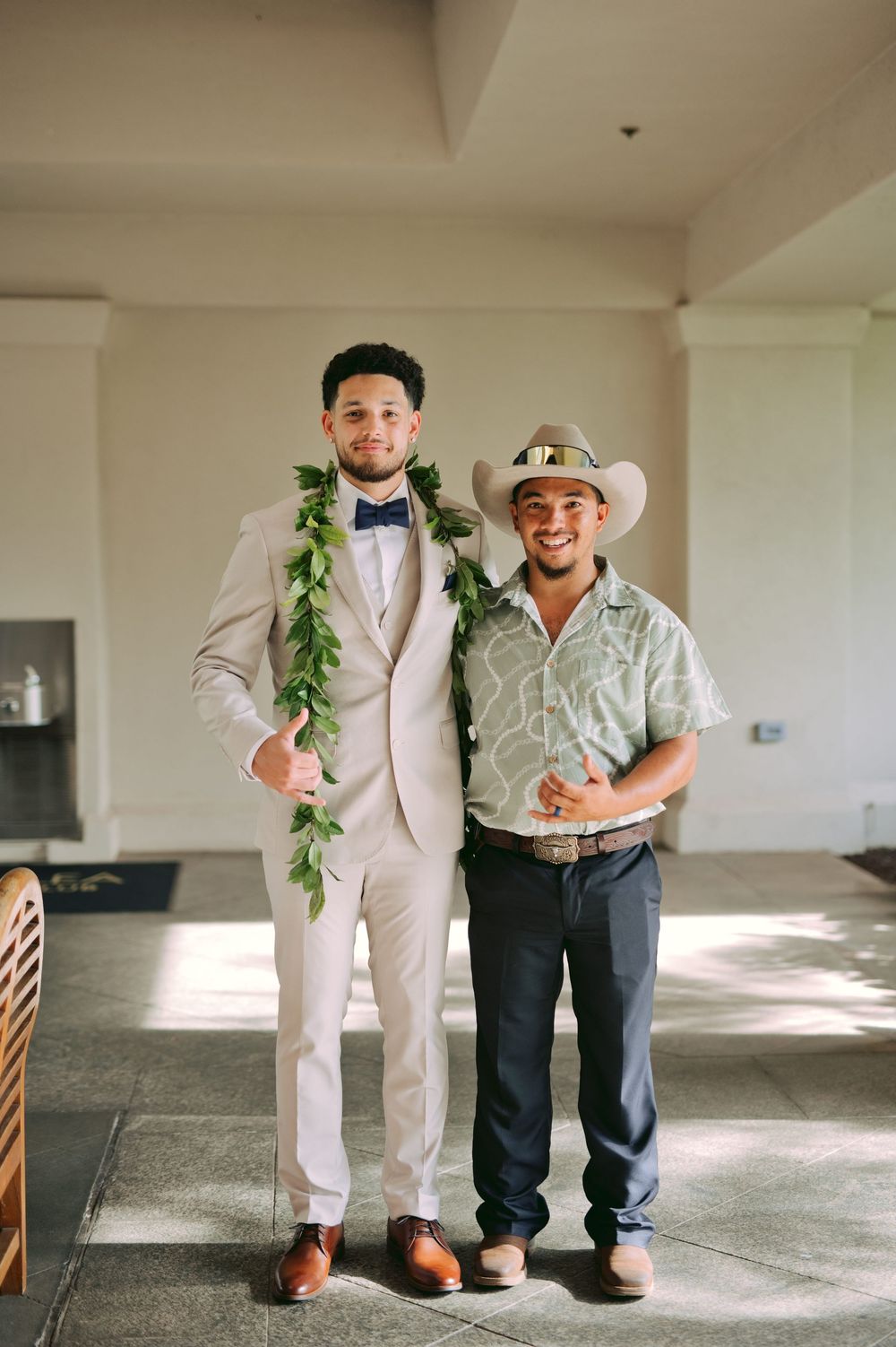 Two people pose together at a formal event, one wearing a light suit with a leafy lei and the other in western attire.