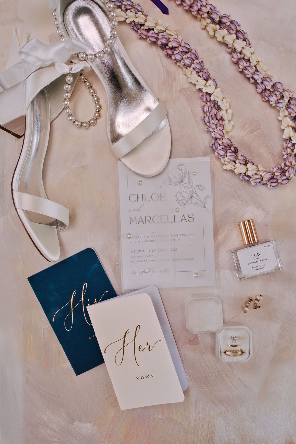 Wedding details including invitations, jewelry, and perfume bottles arranged on a neutral textured surface.
