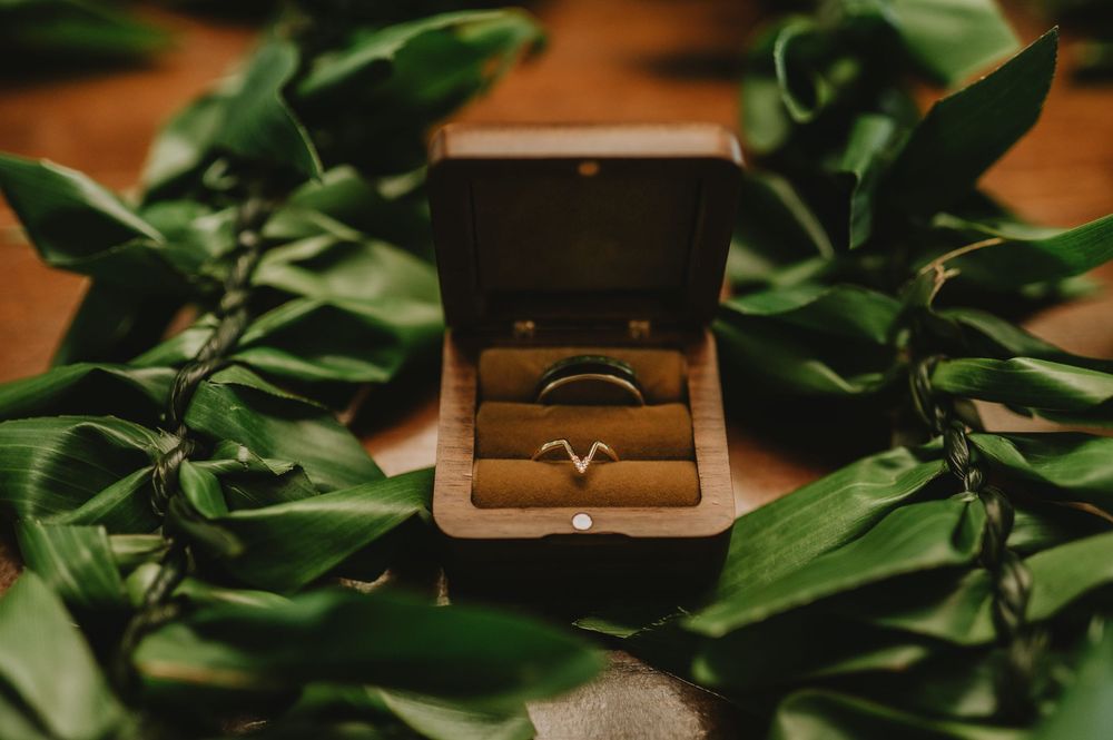 Wedding rings rest in wooden box surrounded by lush green tropical leaves on wooden surface.