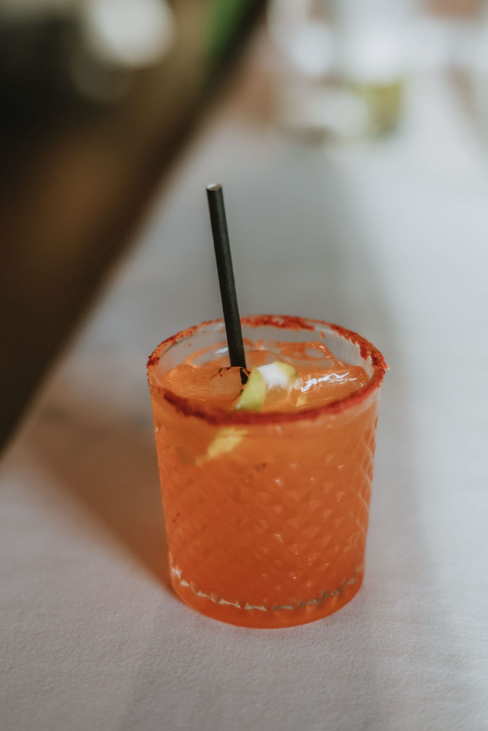 Row of orange cocktails with salt rims and black straws served on white tablecloth.