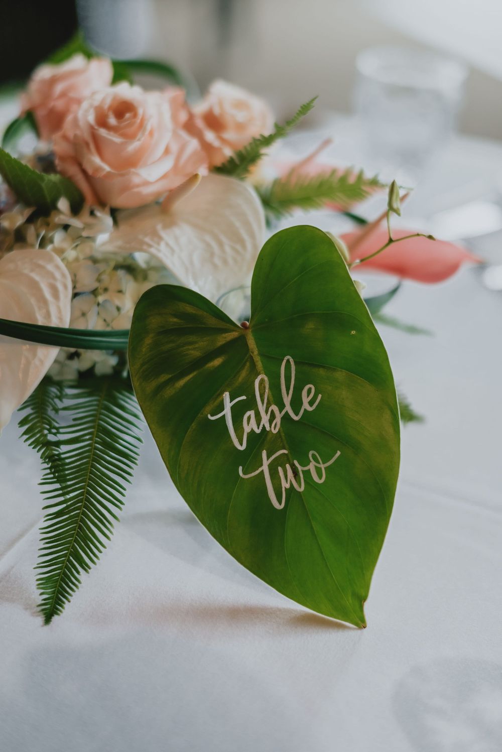 Wedding table number 'two' written in white calligraphy on a tropical green leaf with peach roses and ferns in background.