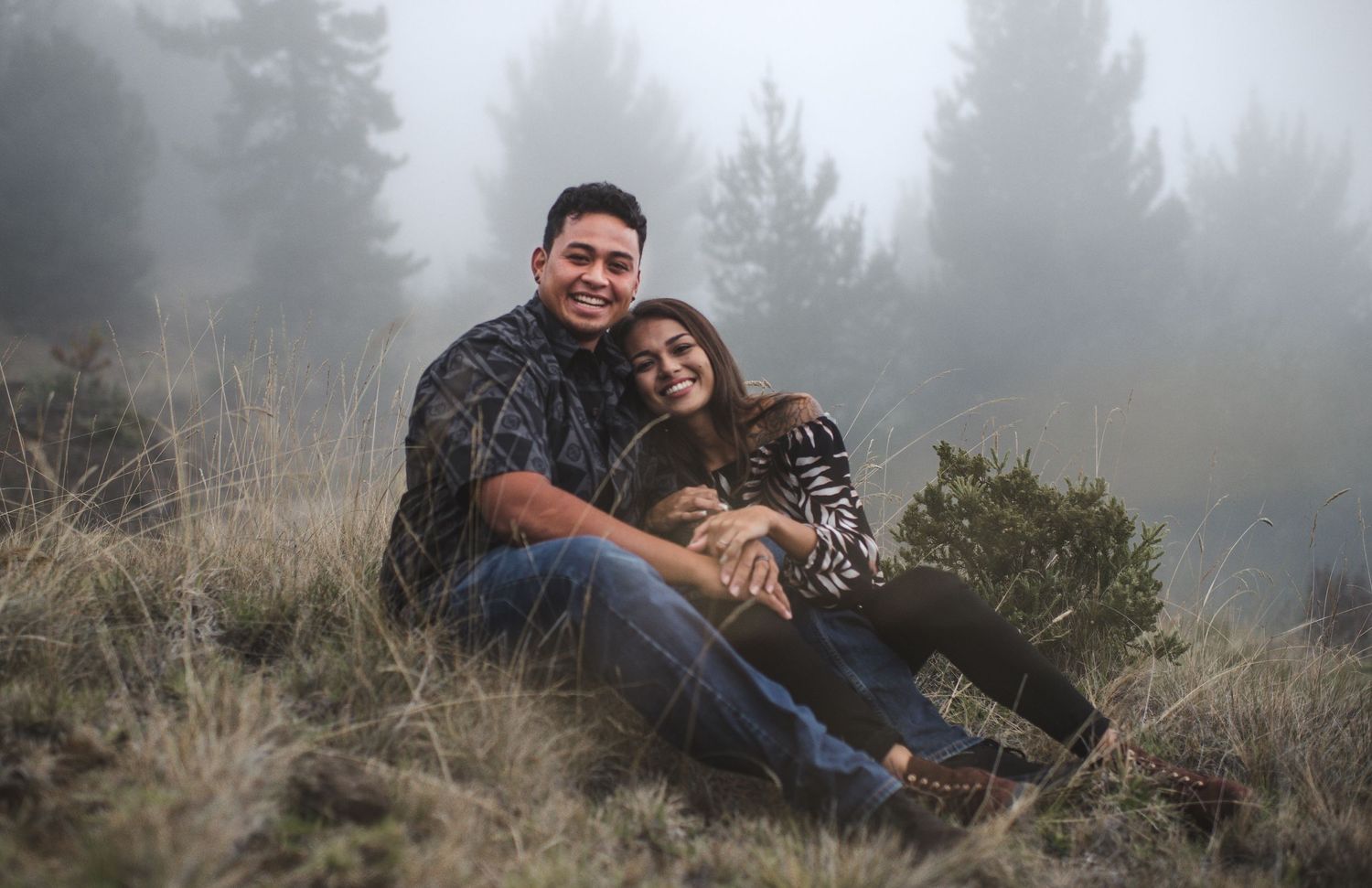 Couple relaxes together on a foggy hillside with pine trees in the background.
