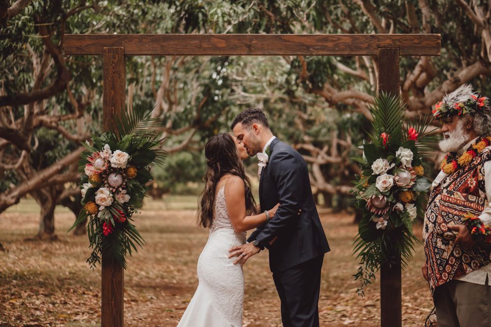 Wedding ceremony in progress under a wooden arch decorated with tropical flowers and greenery.