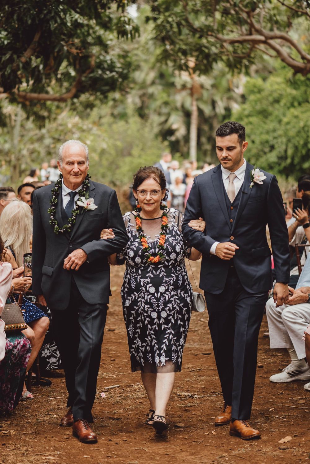 Wedding guests walk in a playful line through a wooded outdoor venue.