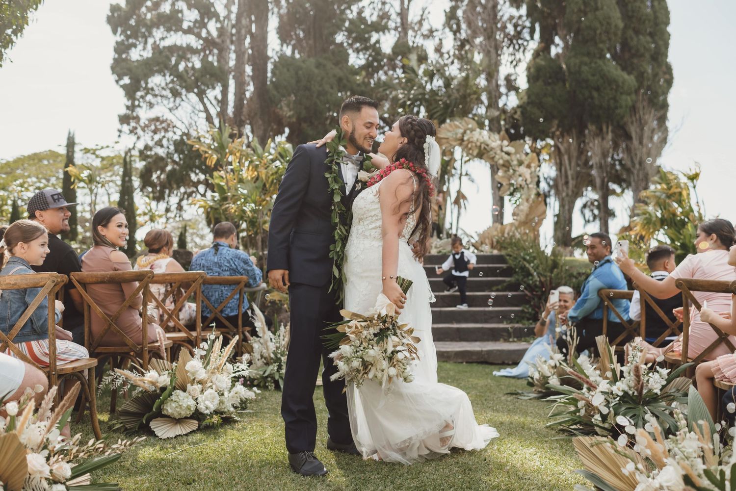 Outdoor wedding ceremony setup with white chairs and floral arrangements under tall trees.