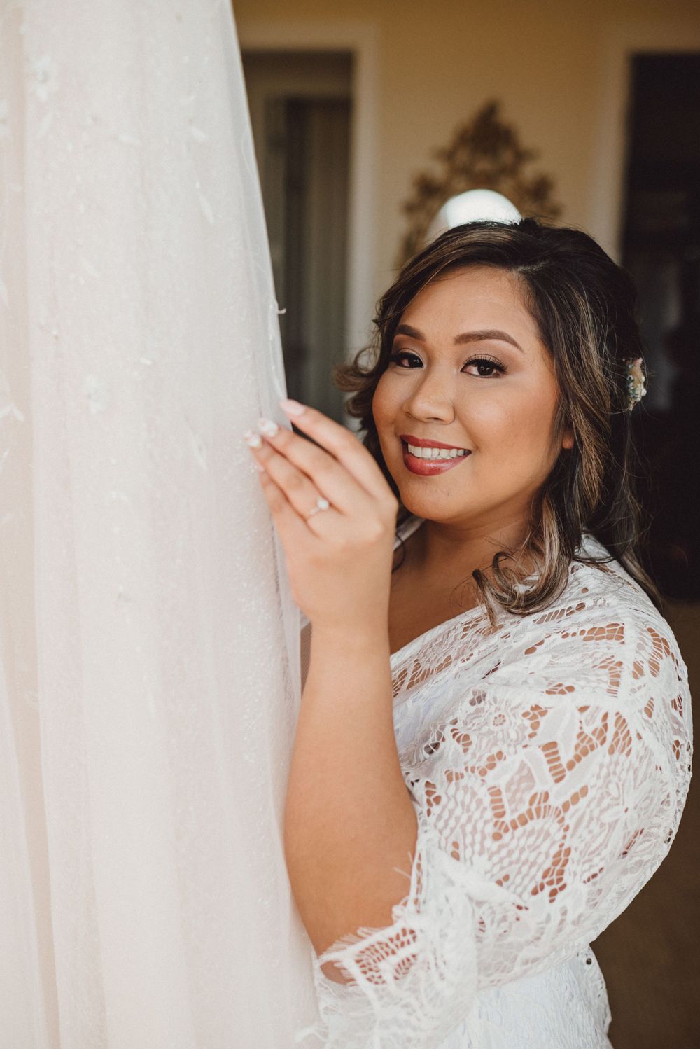 Bride in white lace robe poses playfully near white curtains.