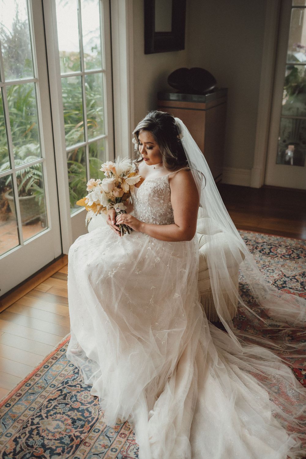 Bride in flowing white gown seated elegantly by large windows with natural light.