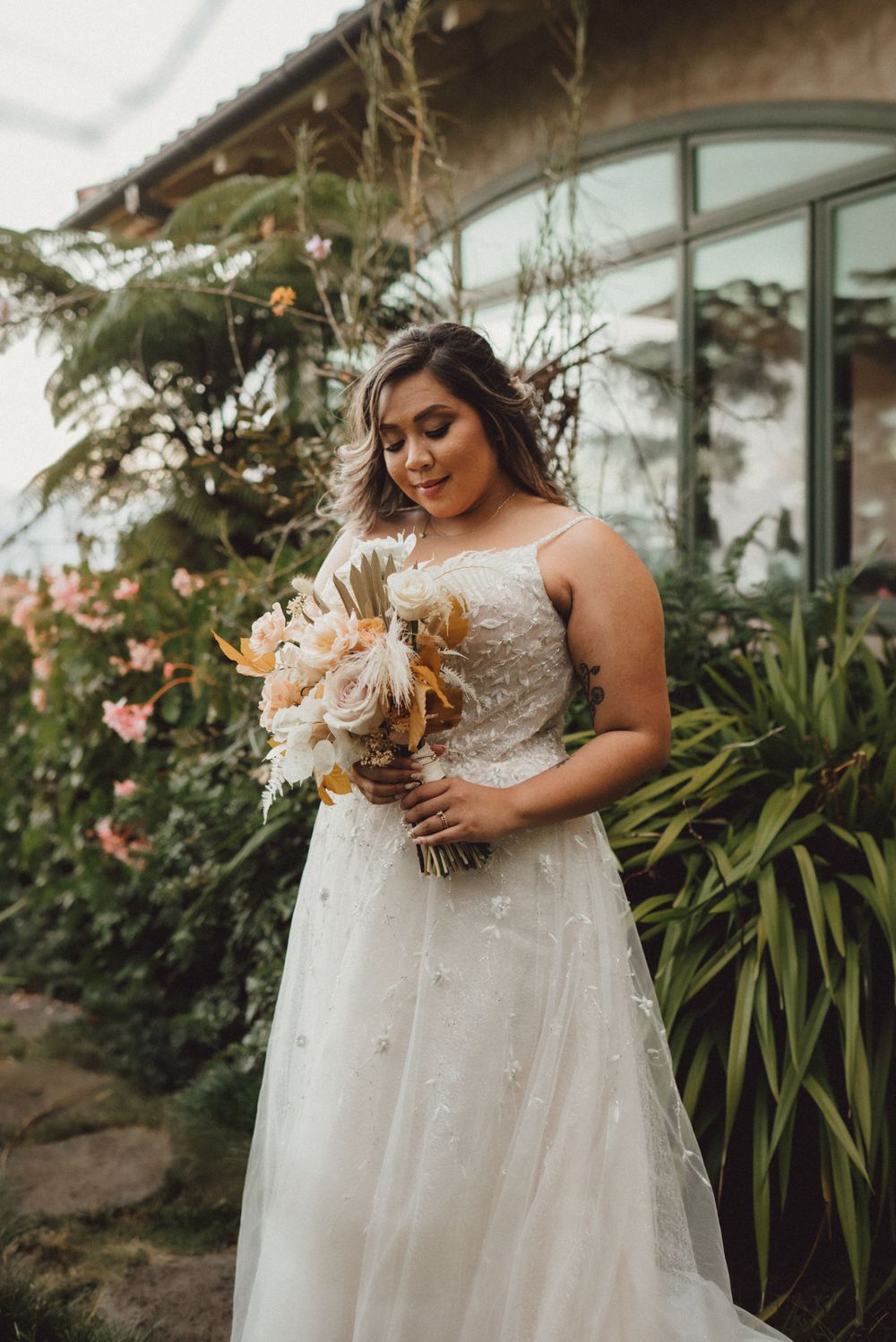 A bride in a lace wedding dress holds a beautiful bouquet of white and peach flowers in a garden setting.