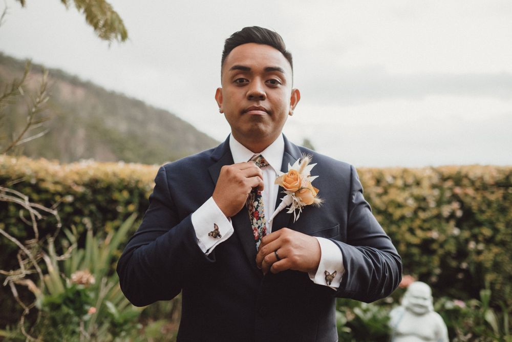A groom adjusts his boutonniere while wearing a classic navy suit outdoors.