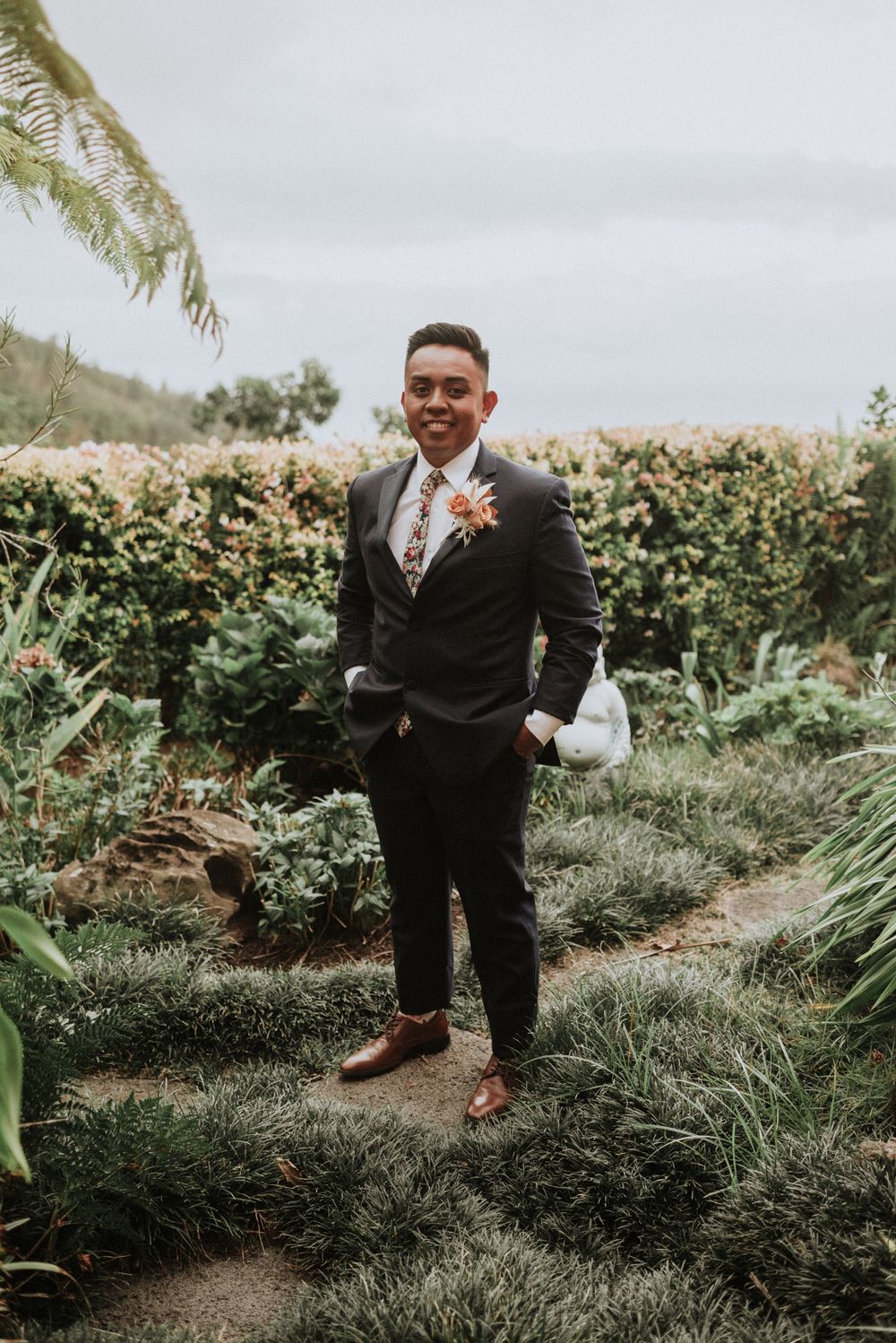 A groom in a dark suit smiles while standing in a lush garden landscape.