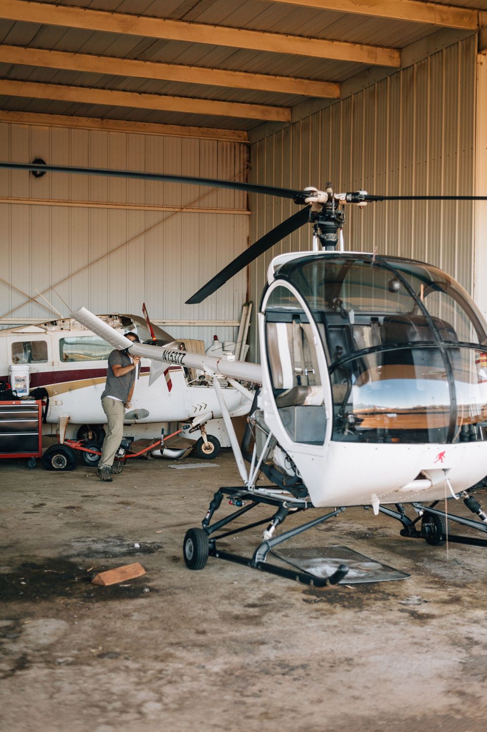 Row of light aircraft stored in a large hangar with exposed wooden ceiling beams.
