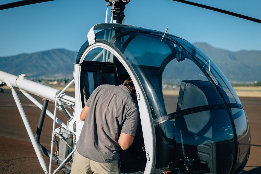 Helicopter pilot inspecting rotor blades against bright blue sky and mountain backdrop.