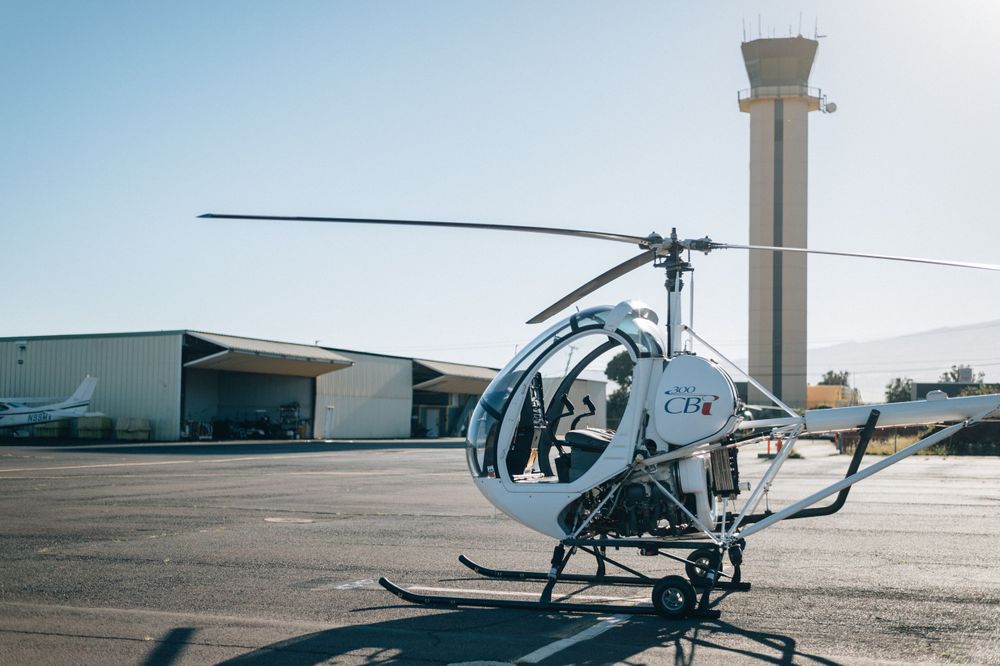 Small white helicopter parked near airport control tower under clear sky.