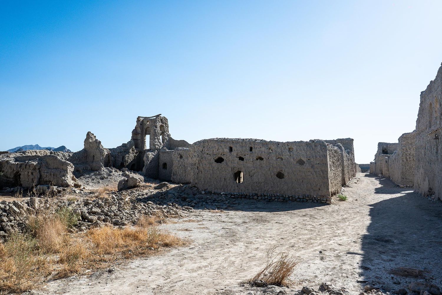 Ancient stone ruins and crumbling walls line a dusty pathway through an archaeological site under bright blue skies.