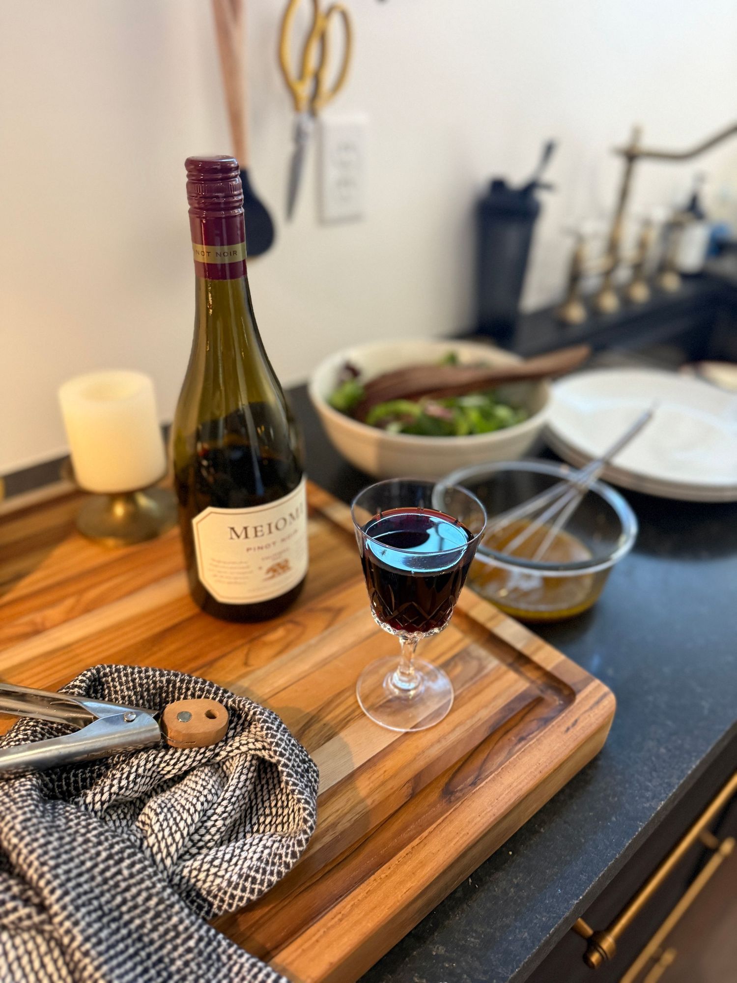 A glass of red wine and bottle sit on a wooden serving board with a black and white patterned napkin.