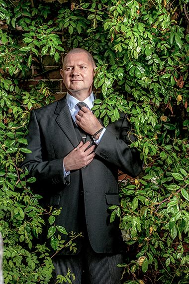 Professional portrait of someone in a dark business suit adjusting their tie while standing against a leafy green wall.