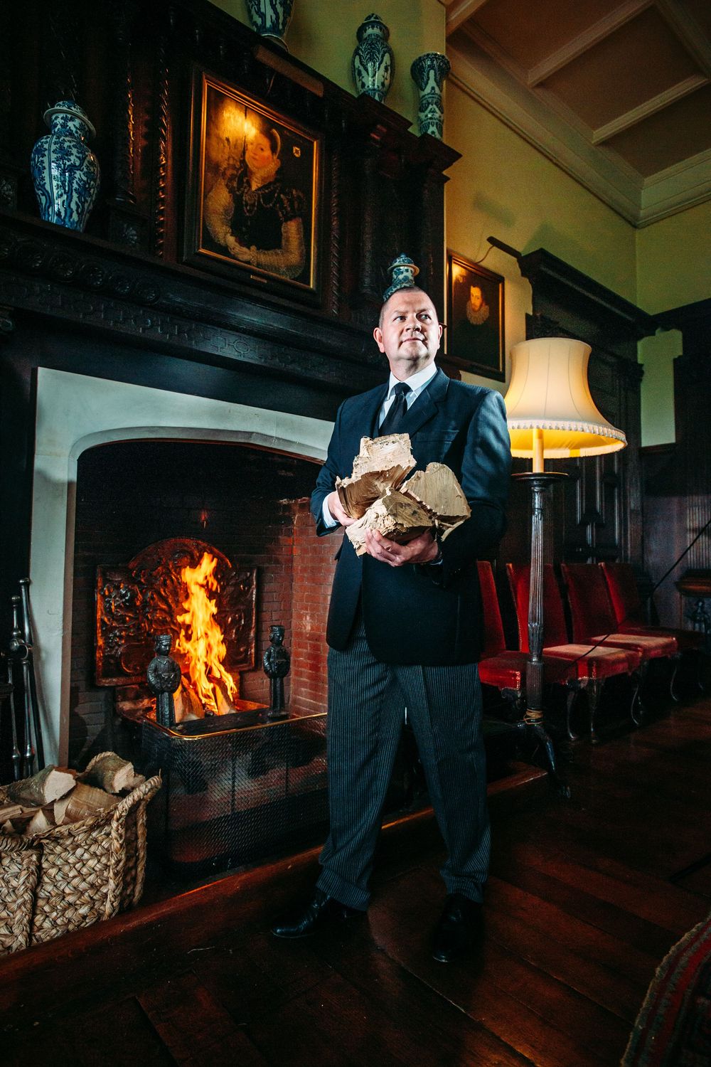 A person in formal attire tends to a rustic stone fireplace in a dimly lit room with wooden floors and vintage decor.