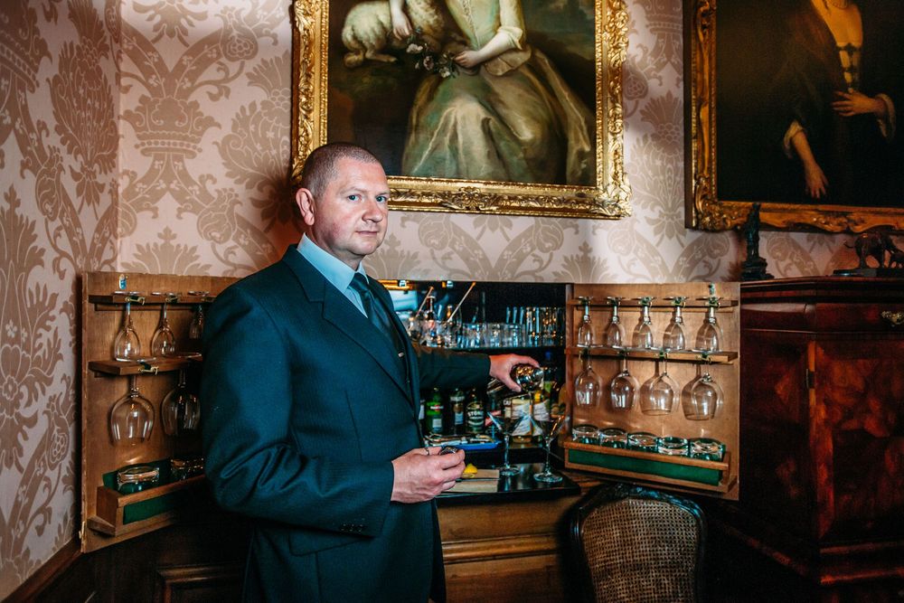 A person in a dark suit looks at their phone while standing in an ornate room with vintage wallpaper and paintings.